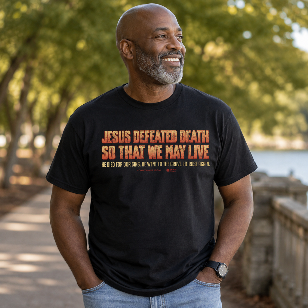 A smiling man wearing a black graphic t-shirt with orange and white text that reads 'JESUS DEFEATED DEATH SO THAT WE MAY LIVE' and 'HE DIED FOR OUR SINS, HE WENT TO THE GROVE, HE ROSE AGAIN.' He is standing outdoors on a sunny path with trees and water in the background.