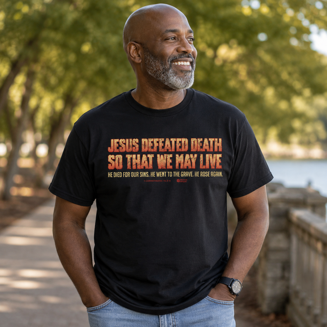 A smiling man wearing a black graphic t-shirt with orange and white text that reads 'JESUS DEFEATED DEATH SO THAT WE MAY LIVE' and 'HE DIED FOR OUR SINS, HE WENT TO THE GROVE, HE ROSE AGAIN.' He is standing outdoors on a sunny path with trees and water in the background.