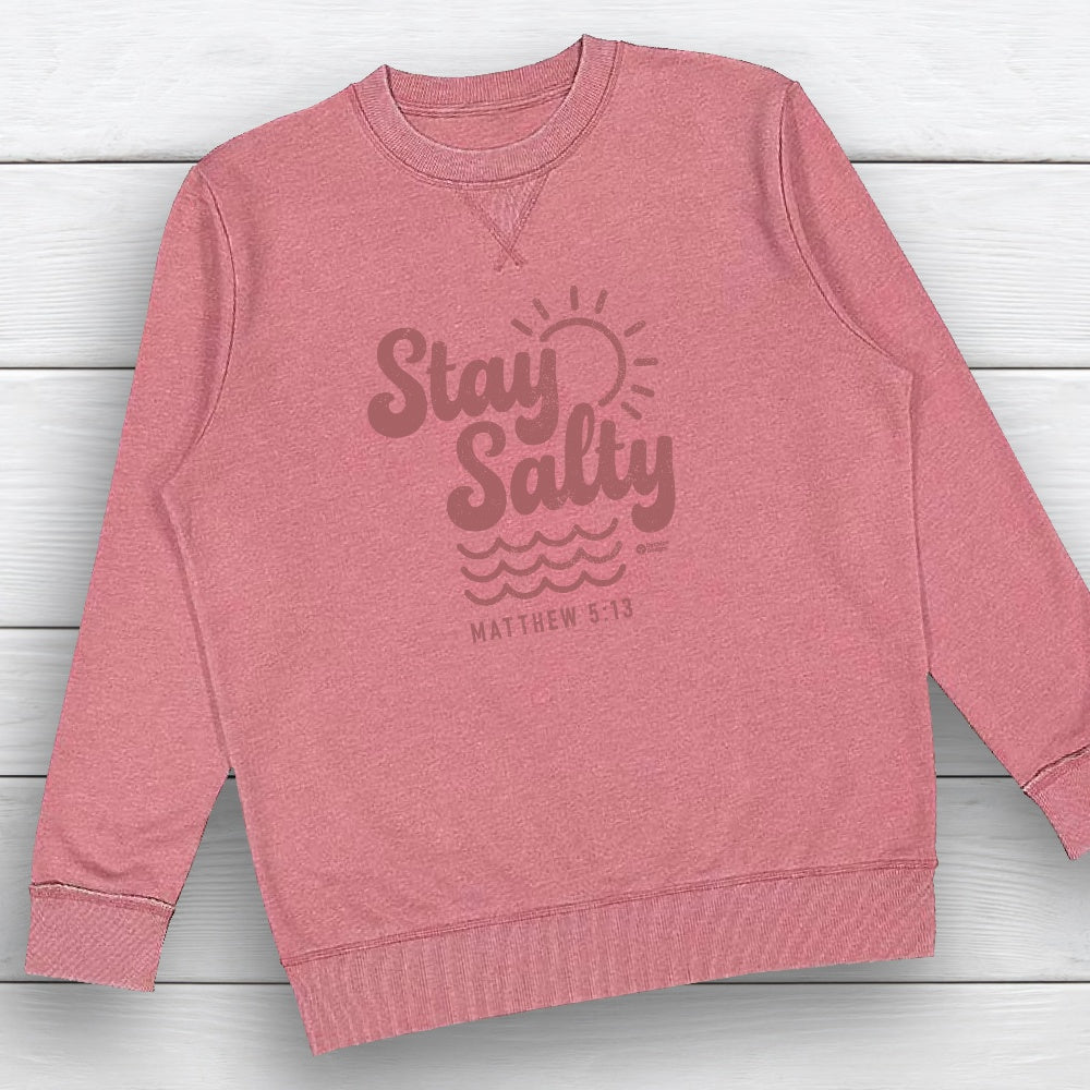 A heathered pink crewneck sweatshirt with a dark pink graphic on the front that reads 'Stay Salty' with a sun and waves, and 'MATTHEW 5:13' below, laid flat on a white wood plank background.
