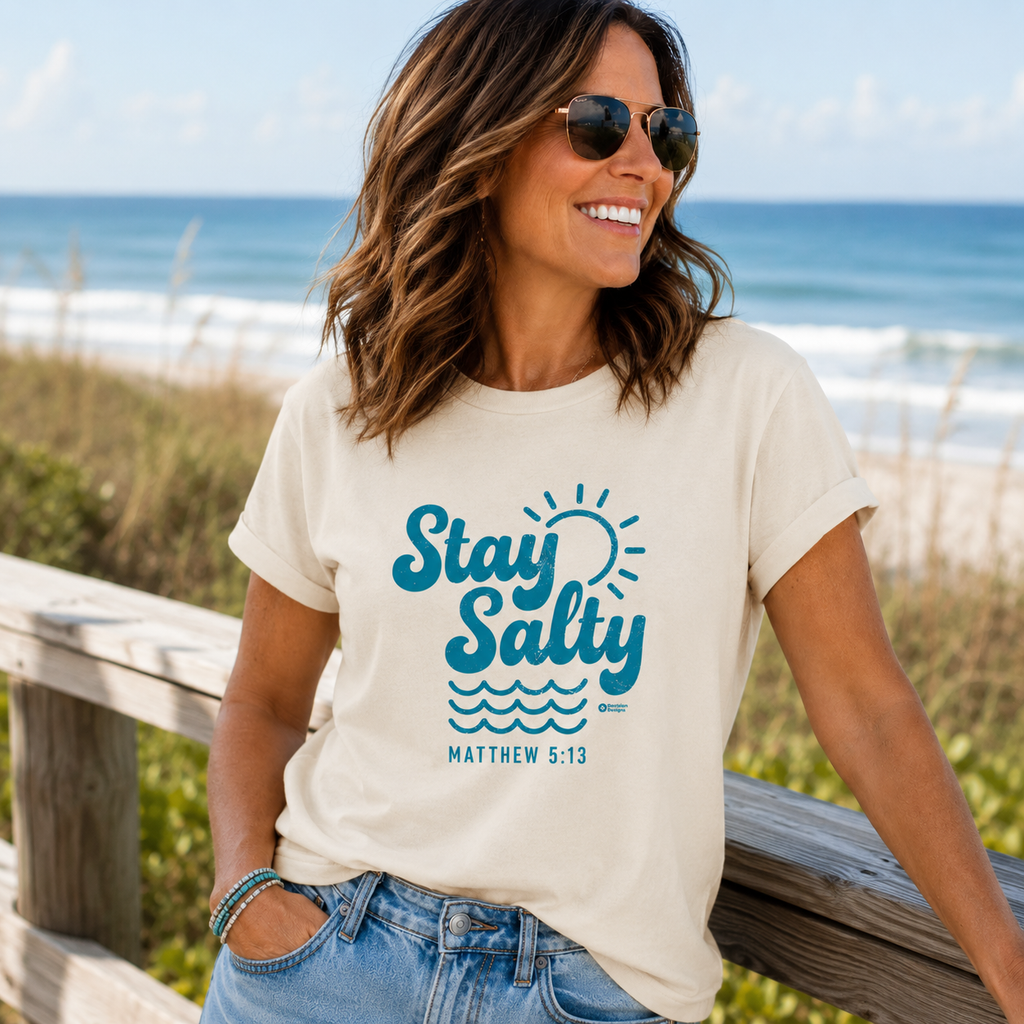 A woman wearing a cream-colored graphic t-shirt with a blue 'Stay Salty' design and 'MATTHEW 5:13' text, standing on a wooden boardwalk with the ocean in the background.