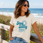 A woman wearing a cream-colored graphic t-shirt with a blue 'Stay Salty' design and 'MATTHEW 5:13' text, standing on a wooden boardwalk with the ocean in the background.